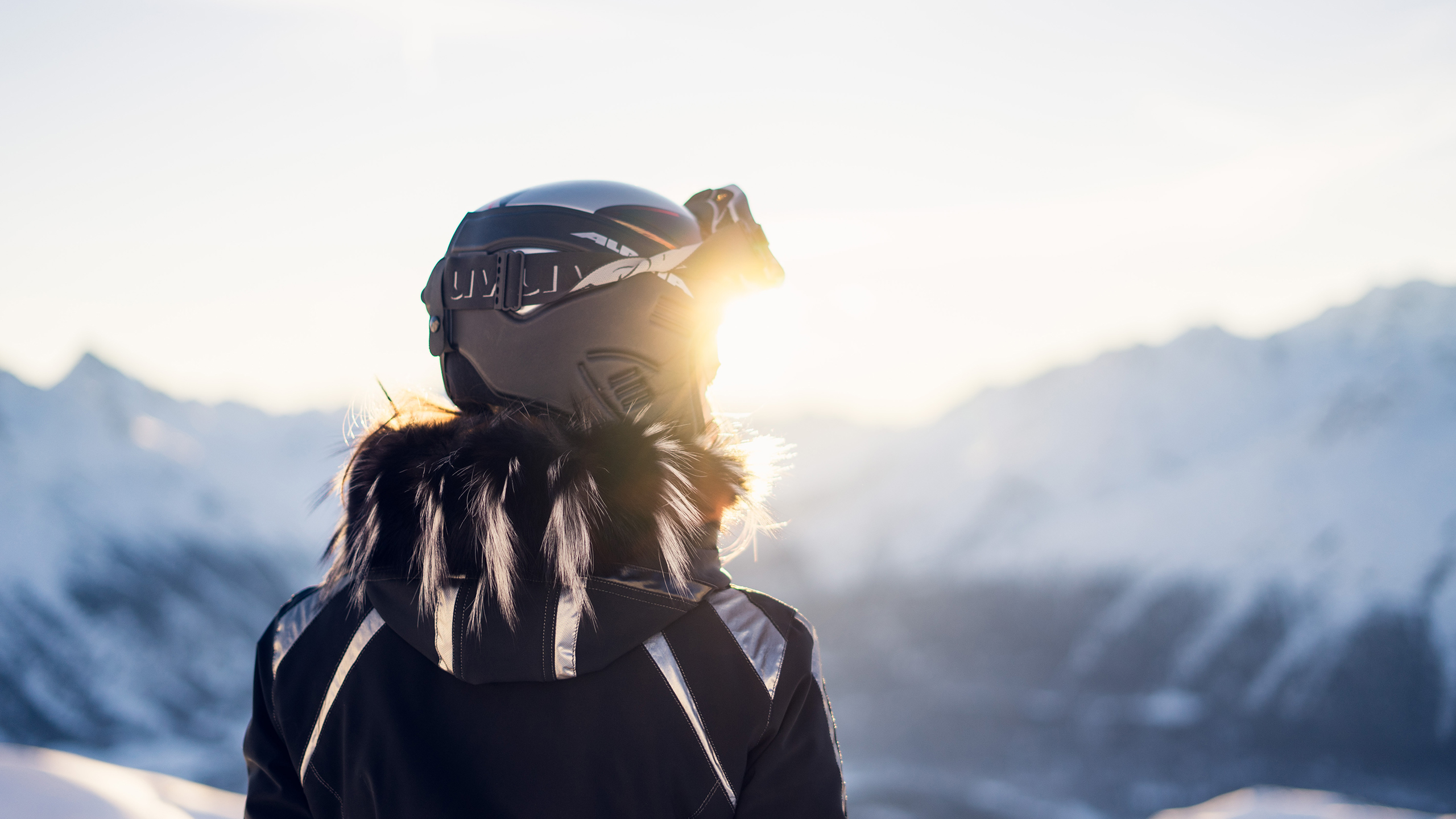 Une femme de dos est en train de regarder l'horizon sur une piste de ski. Elle est équipée d'un casque et de lunettes de ski