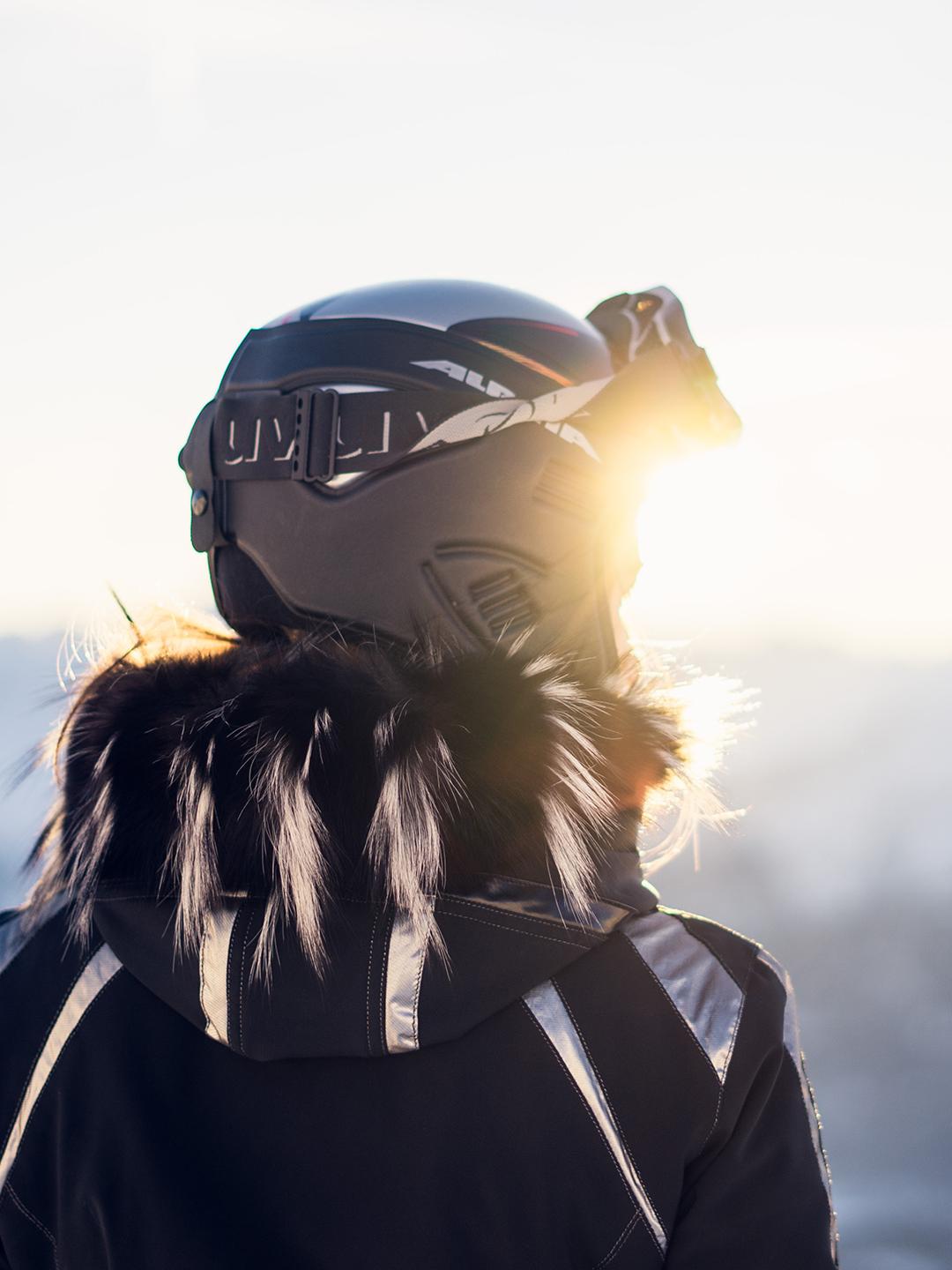 Une femme de dos est en train de regarder l'horizon sur une piste de ski. Elle est équipée d'un casque et de lunettes de ski