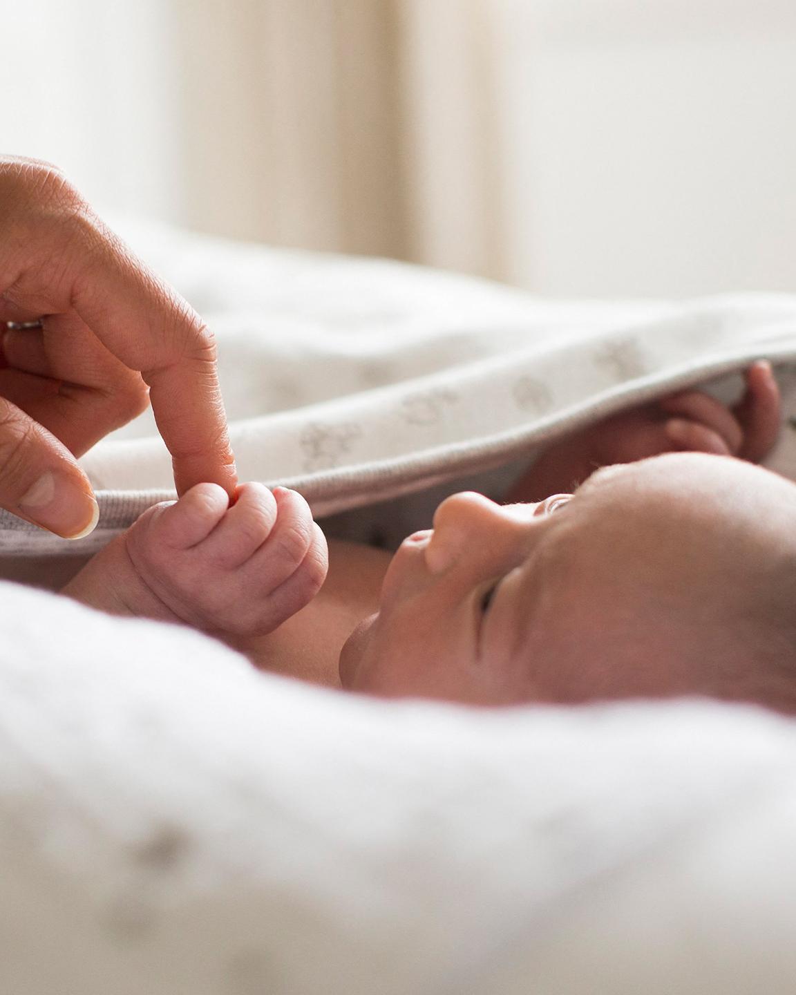 Un bébé est allongé sur un matelas avec un drap sur lui, il tient dans sa main le doigt d'une personne