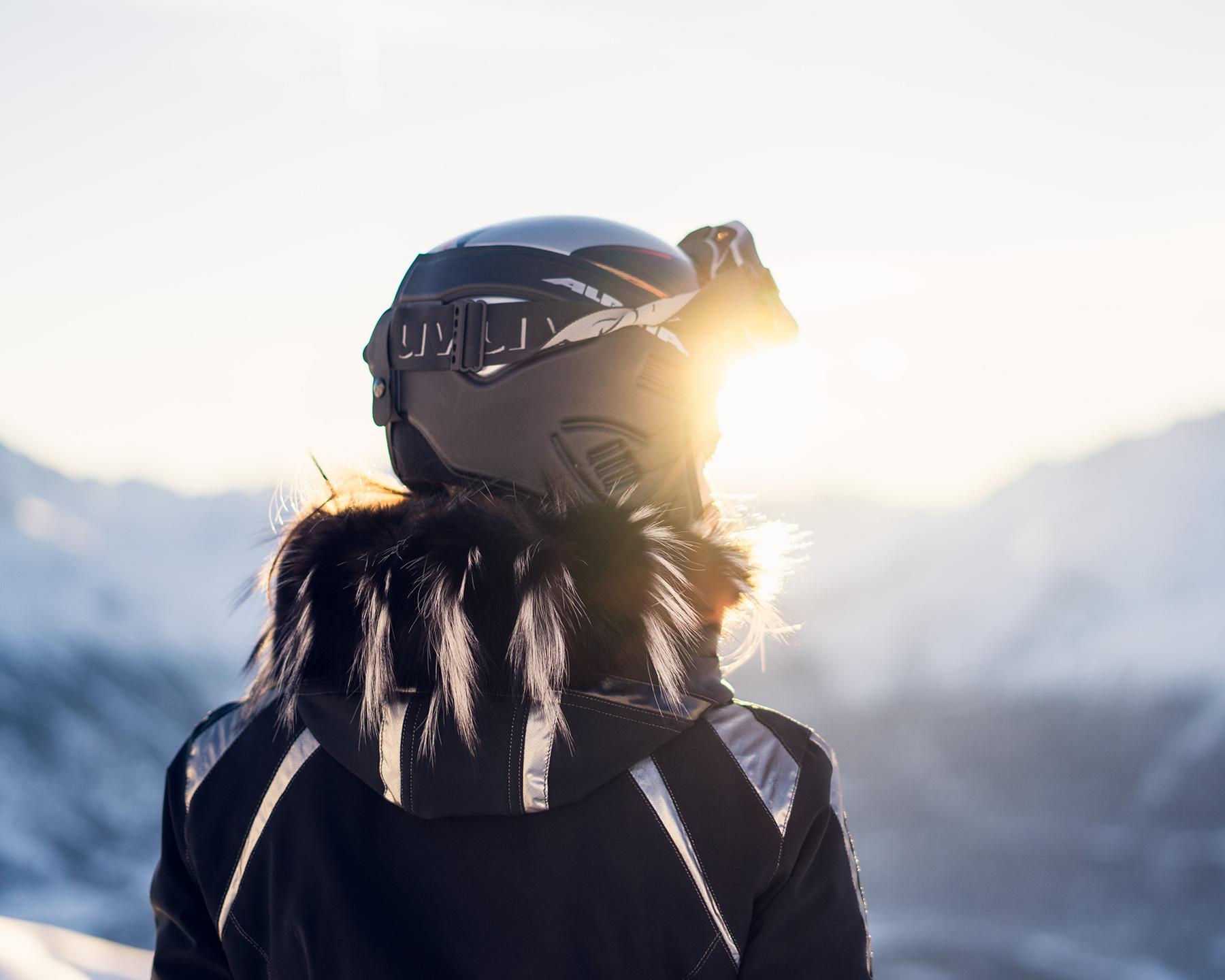 Une femme de dos est en train de regarder l'horizon sur une piste de ski. Elle est équipée d'un casque et de lunettes de ski